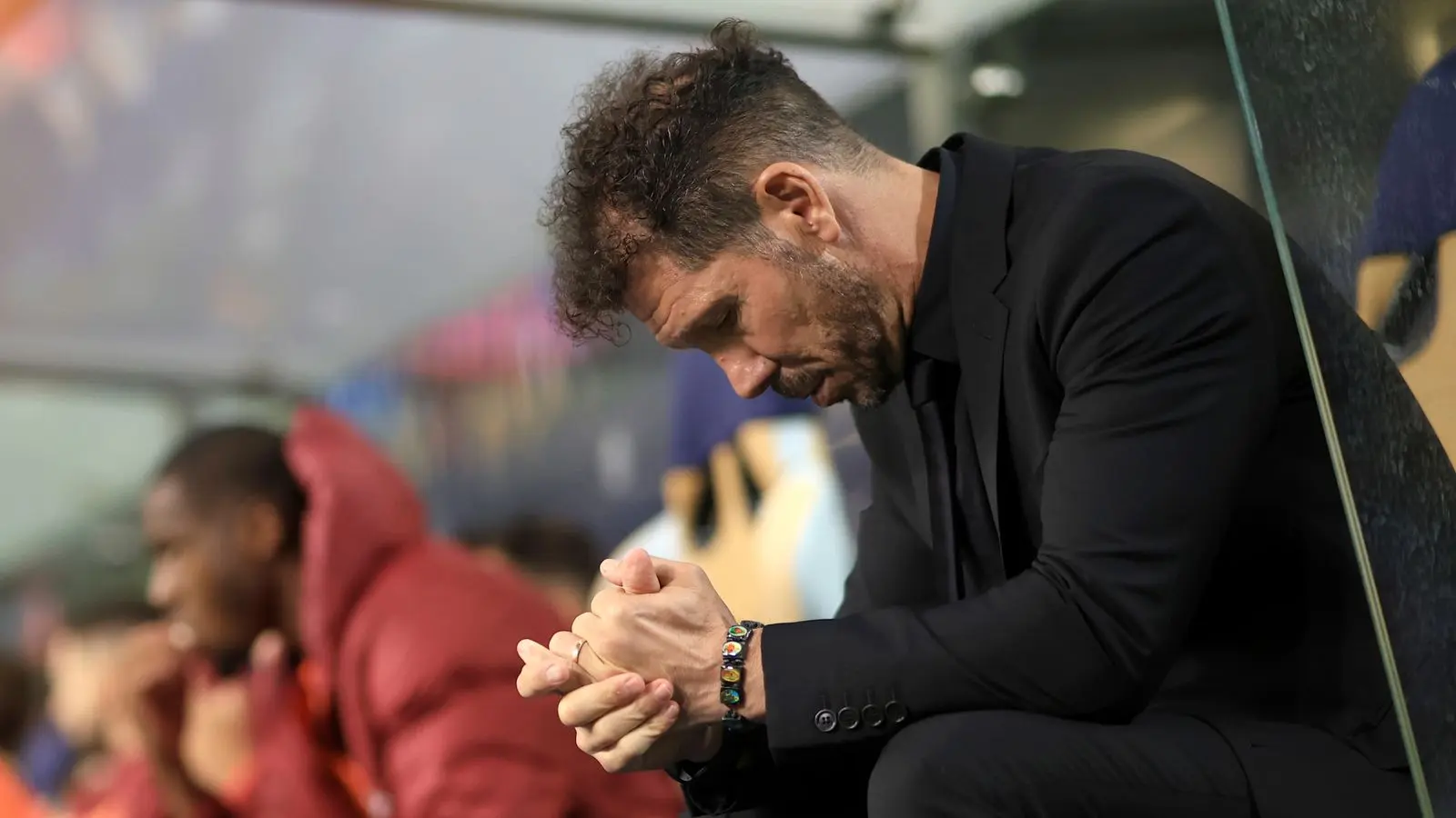 Diego Simeone in a black suit sitting on the Atletico Madrid sideline with his hands clasped.