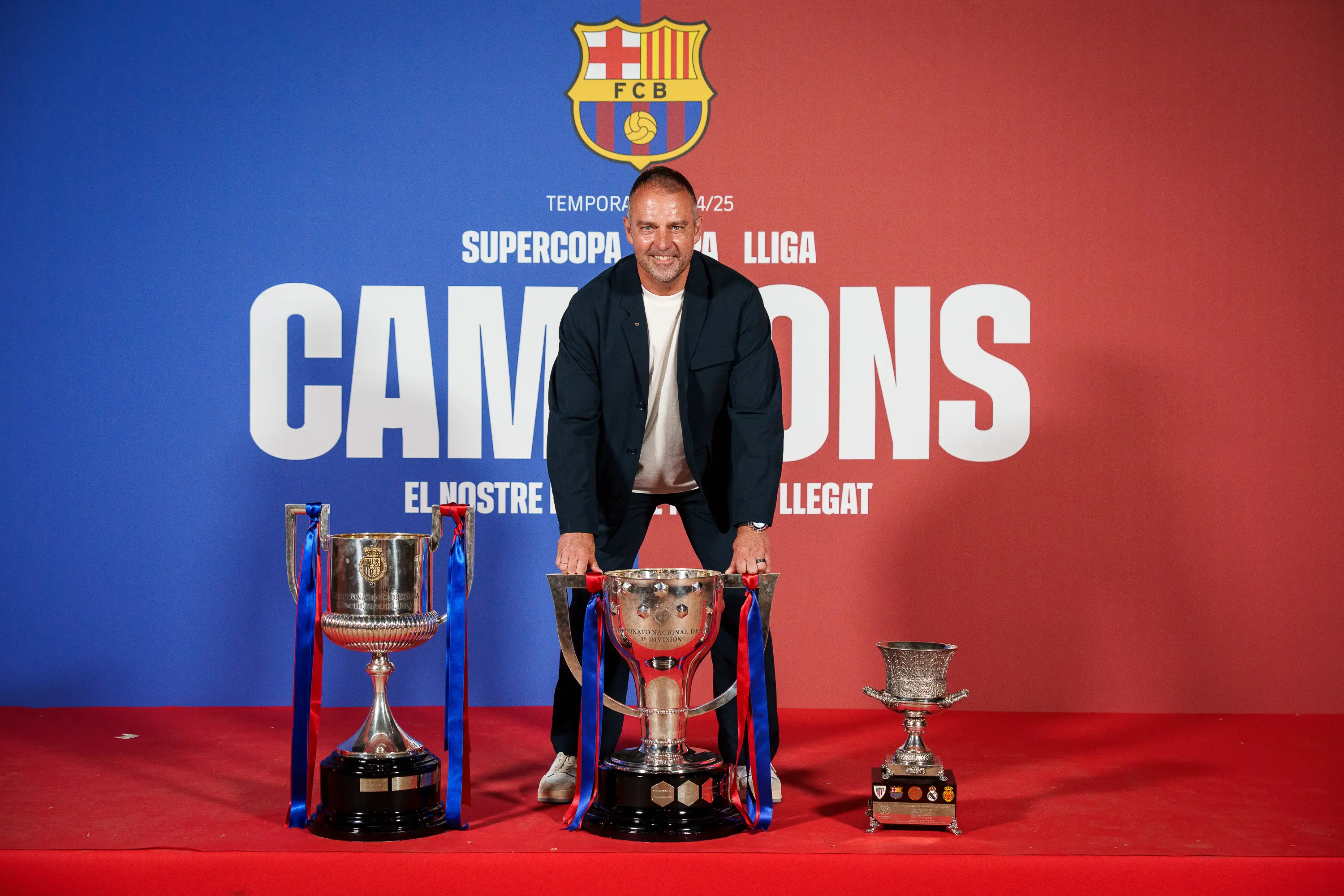 Barça manager Hansi Flick poses with trophies on a red carpet backdrop.