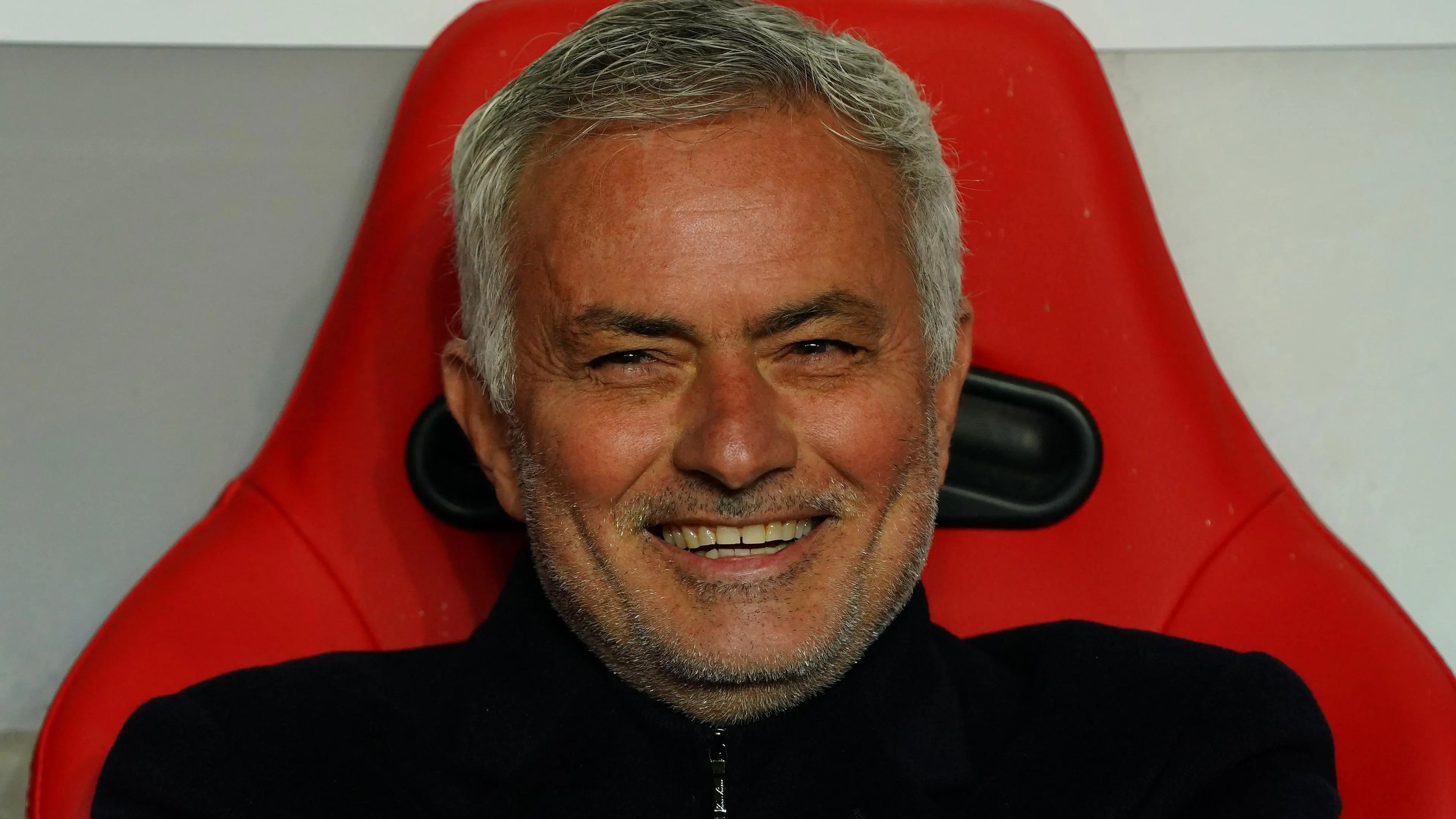 Jose Mourinho smiling while seated in a red coach's seat.