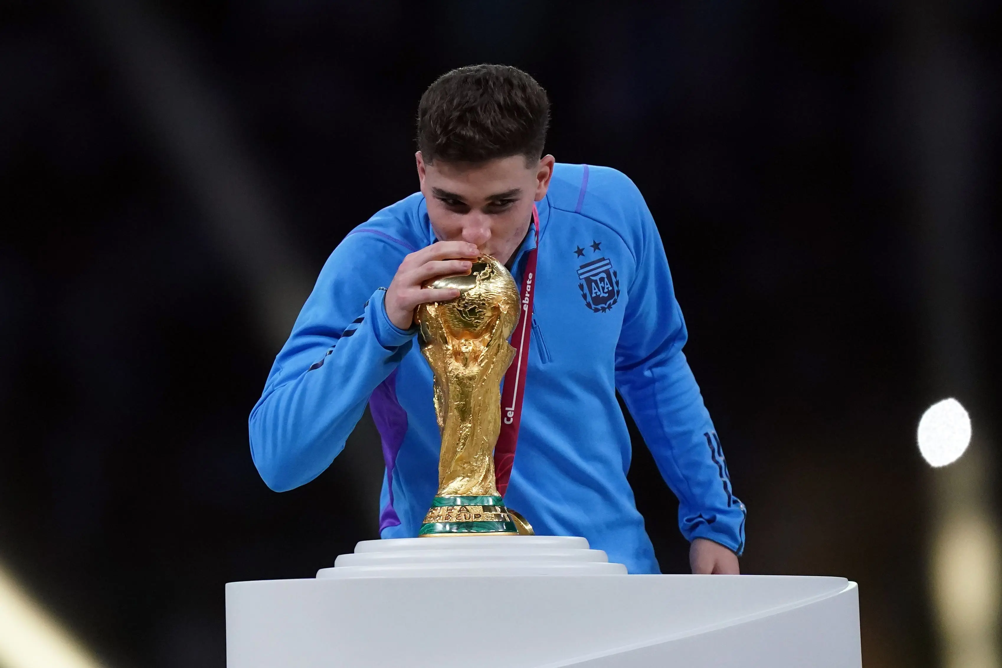 Julian Alvarez kissing the FIFA World Cup trophy, wearing Argentina's blue jersey.