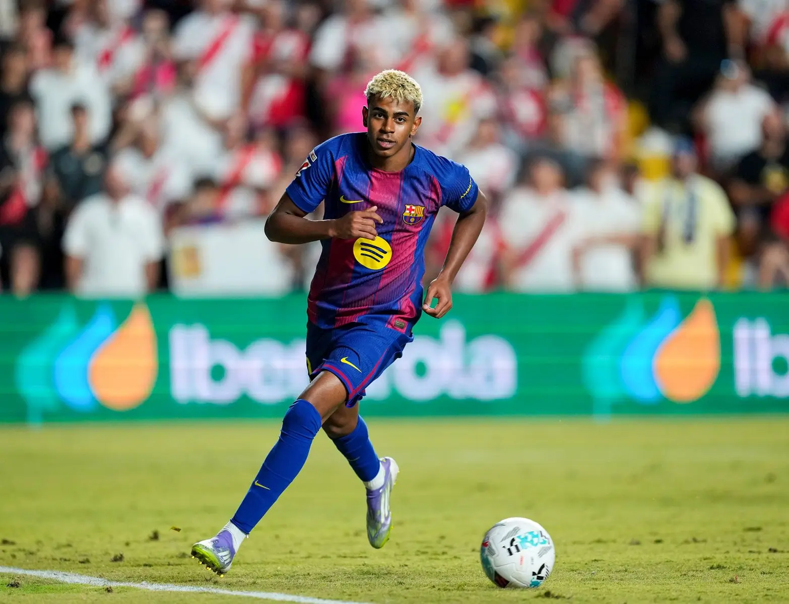 Lamine Yamal in a blue FC Barcelona kit dribbling a soccer ball on a grass field during a match.