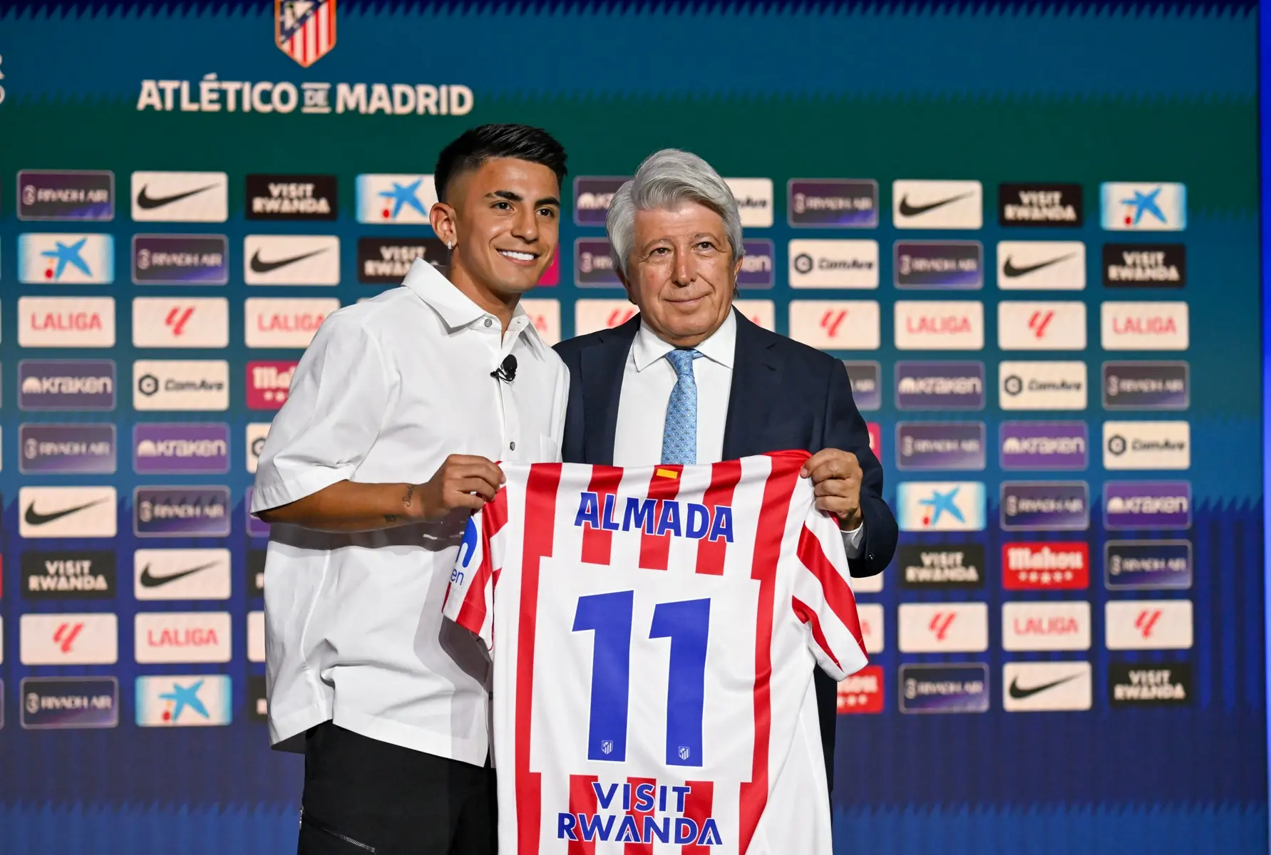 Thiago Almada and an official holding up an Atletico Madrid jersey with Almada's name and number 11.