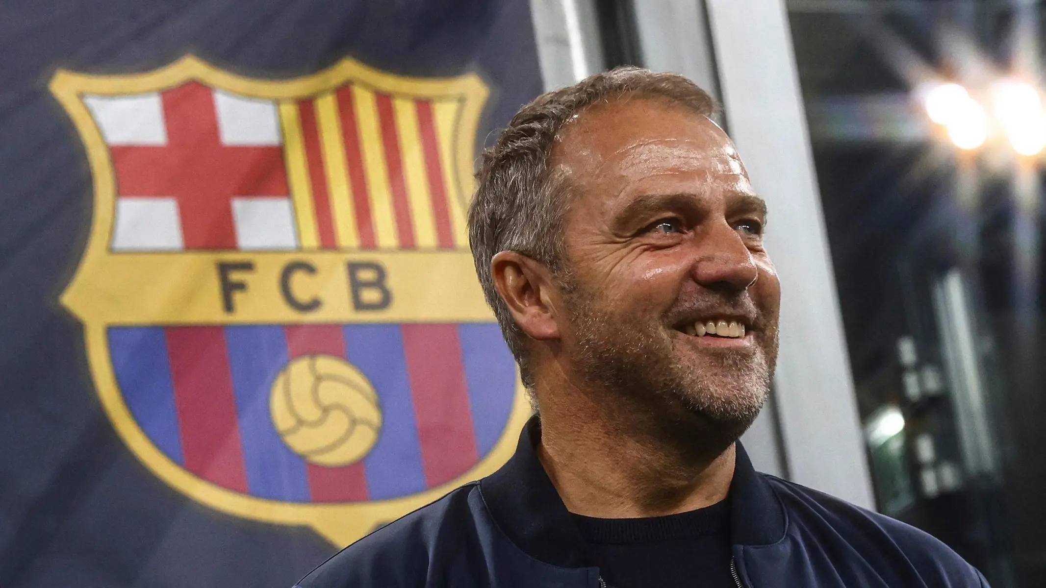 Hansi Flick smiling in front of the FC Barcelona club crest