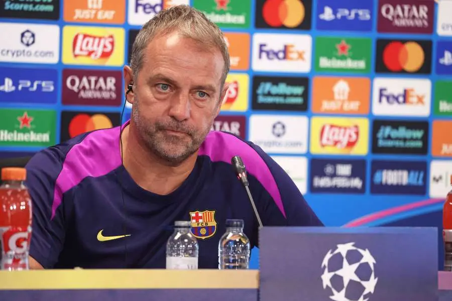 Hansi Flick wearing an FC Barcelona training shirt at a press conference with a sponsor backdrop.