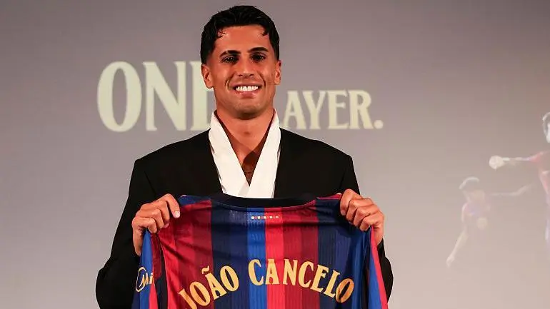 Joao Cancelo wearing a black suit and holding up a Barcelona jersey with his name on the back