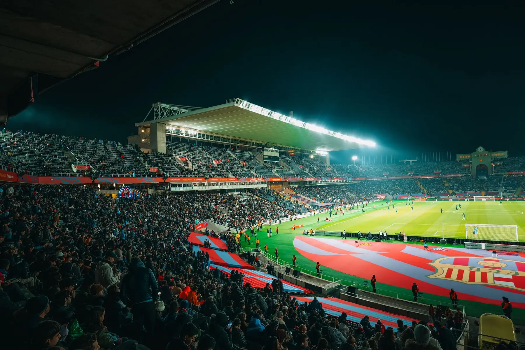 Wide shot of Camp Nou stadium at night with a crowd of fans and the FC Barcelona logo on the pitch
