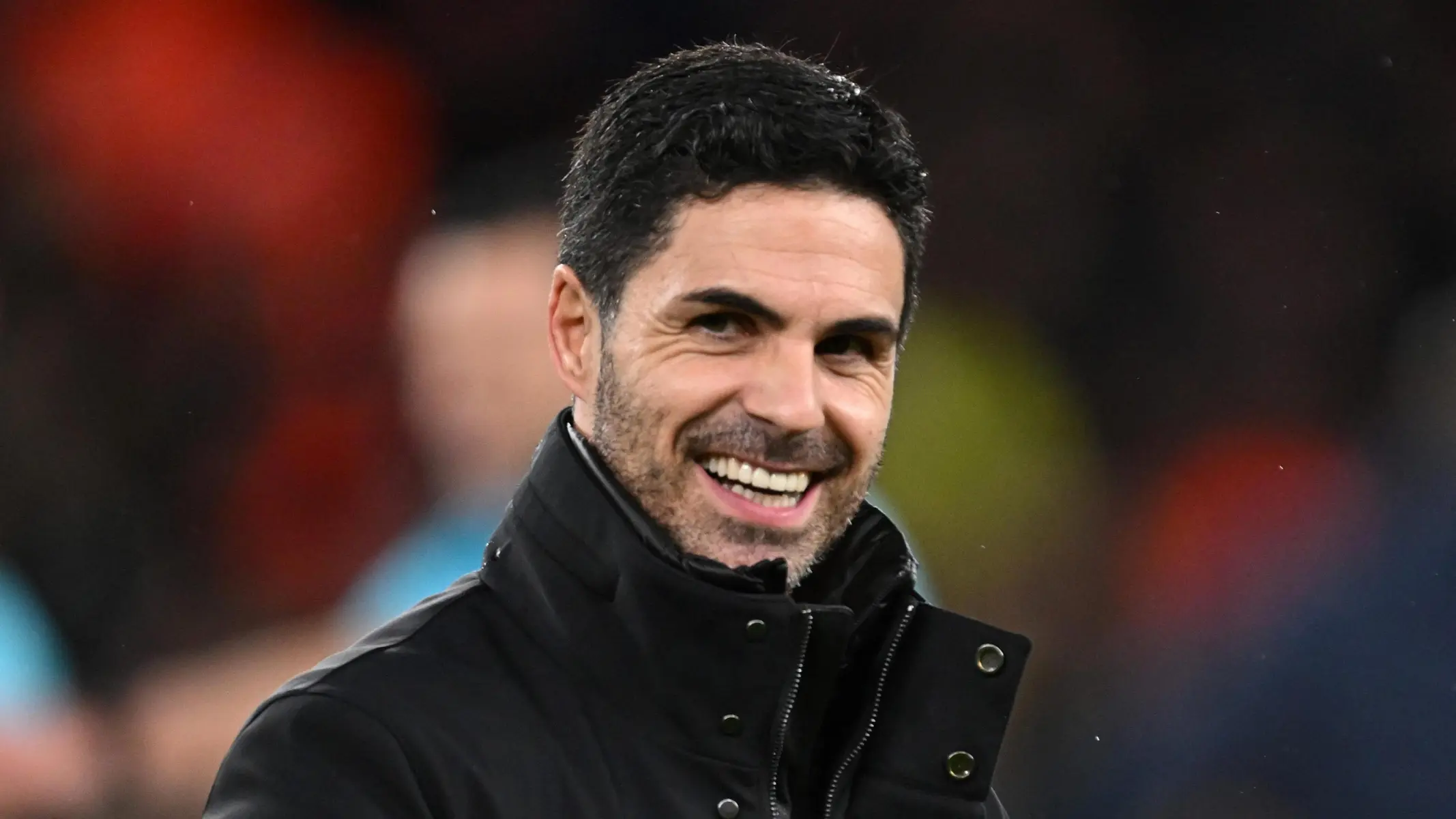 Portrait of Mikel Arteta smiling, wearing a black jacket against a blurred stadium background.