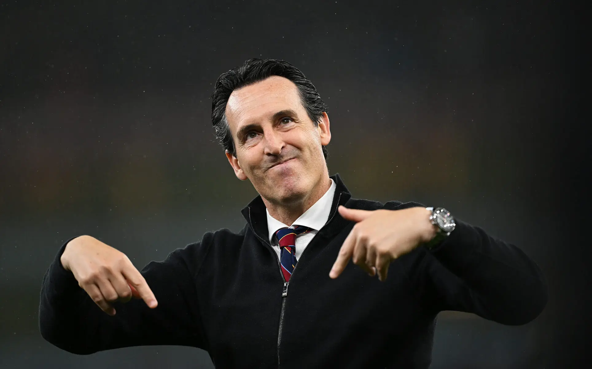 Unai Emery wearing a black jacket and tie, pointing downwards with both hands and smiling.