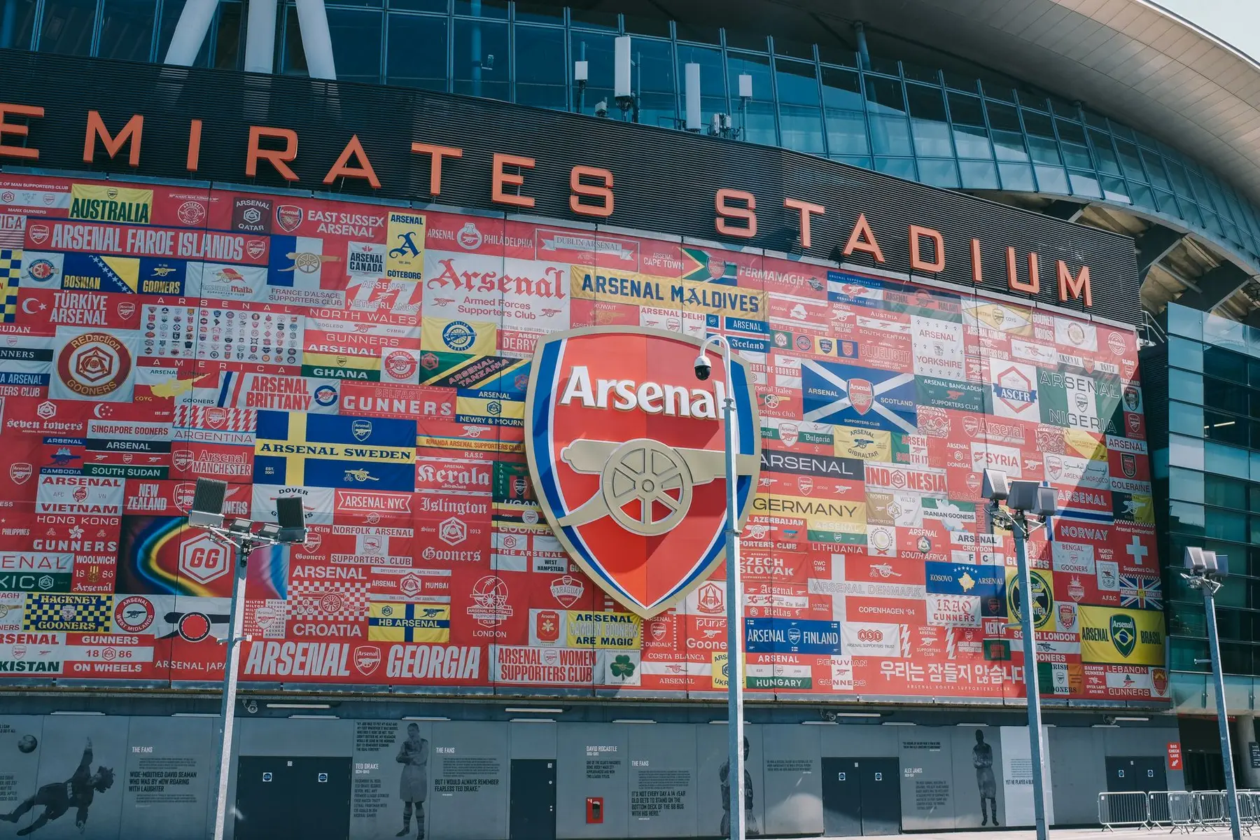 Exterior of Emirates Stadium featuring the Arsenal FC logo and a wall of international fan club banners.